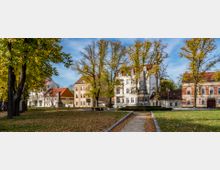 Stadtpark mit Baumreihen, gefallenen Laubblättern und einem zentralen Denkmal in Obeliskform; dahinter stehen historische Gebäude mit roten und hellen Dächern.