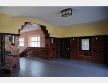 Innenansicht eines Treppenhauses mit Jugendstil-Elementen, geprägt von glasierten braunen Wandfliesen, schwarzen Ornamenten und gelben Wänden. Ein Bogen führt zu einem Treppenbereich mit großen Fenstern; rechts befindet sich eine grüne Tür und eine Informationstafel.