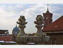 Steinerne, barocke Verzierungen in Form von Vasen mit skulpturalen Köpfen und Ornamenten im Vordergrund, im Hintergrund die Kuppel des Berliner Doms und der Rote Turm des Berliner Rathauses mit Uhr und Fahne.