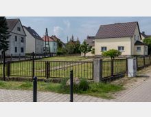 Wohnsiedlung mit Einfamilienhäusern, umgeben von Gärten mit niedrigen Metallzäunen. Im Vordergrund ist ein gepflasterter Gehweg und ein Rasenstück mit einem Stein sowie einem Pfosten zu sehen.