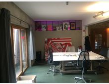 Ein moderner Workshop-Raum mit einem großen Tisch in der Mitte, umgeben von schwarzen Bürostühlen. Im Hintergrund befinden sich eine Wand mit abstrakten Kunstwerken sowie eine Glastür und ein Fenster mit grauen Vorhängen, die einen Holzrahmen haben.