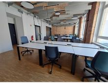 Ein moderner Büroraum mit mehreren weißen Schreibtischen und ergonomischen Bürostühlen auf hellem Holzboden. Die Backsteinwände und die große Fensterfront sorgen für viel Tageslicht, während abgehängte Akustikpaneele und runde Deckenleuchten das Design ergänzen.