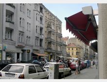 Straßenszene in einer Stadt mit mehrstöckigen Wohnhäusern im Gründerzeitstil, Balkonen und verzierten Fassaden; im Vordergrund geparkte Autos, Fußgänger und ein rotes Markisendach auf der rechten Seite.