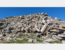 Ein großer Schutthaufen aus zerbrochenen Betonstücken und Steinen, teils bewachsen mit Gras und kleinen Pflanzen, unter einem klaren, blauen Himmel.
