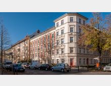 Ecke eines Wohngebäudes im Altbau-Stil mit heller Fassade und roten Akzenten, umgeben von kahlen Bäumen im Herbst. Mehrere geparkte Autos stehen entlang der Straße, und ein Verkehrszeichen weist auf die "Herbert-Baum-Straße" hin.