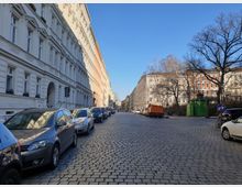 Eine gepflasterte Straßenansicht in einem Wohngebiet mit historischen Gebäuden aus der Gründerzeit, links und rechts gesäumt von geparkten Autos. Rechts ist ein großer, kahler Baum und ein grüner, historischer Kiosk zu sehen, bei klarem, sonnigem Himmel.
