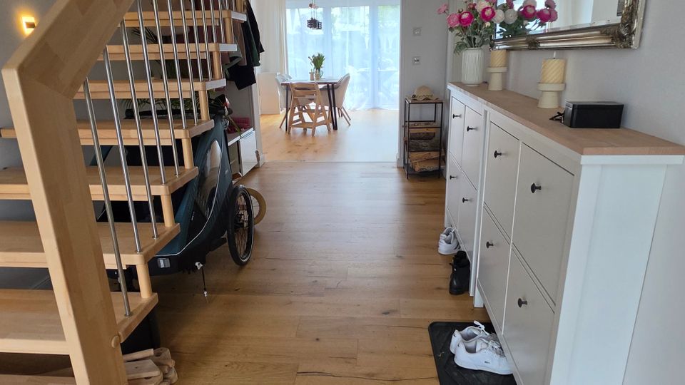 Heller Eingangs- und Flurbereich eines Wohnhauses mit Holztreppe links und einem weißen Sideboard mit großem Wandspiegel rechts. Im Hintergrund führt der Raum zum Essbereich mit Tisch vor einer großen Glas-Schiebetür; Holzfußboden und Blumen-/Girlandendekoration sind sichtbar.