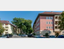 Straßenszene in einer Wohngegend mit mehrstöckigen Wohnhäusern, deren Fassaden beige und rosa gestrichen sind. Im Vordergrund sind parkende Autos und Bäume, die die Straße säumen.