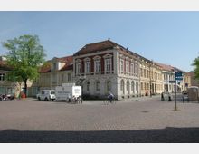 Eine gepflasterte Platzfläche mit umliegenden historischen Gebäuden, darunter ein auffälliges rot-weißes Haus im Stil des Klassizismus mit verzierten Fenstern und Bögen. Im Vordergrund befinden sich Fußgänger, Fahrradfahrer und geparkte Fahrzeuge, darunter ein Lieferwagen mit einem Anhänger.