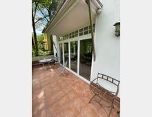 Eine Terrasse mit orangefarbenen Fliesen, vor einer weißen Hausfassade mit großen, bodentiefen Fenstern. Auf der Terrasse stehen weiße Metall-Tische und -Stühle, umgeben von Bäumen und grüner Vegetation im Hintergrund.