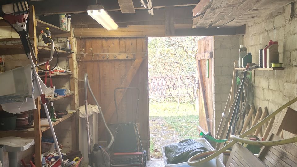 Innenansicht eines Geräteschuppens mit freiliegenden Holzbalken und Neonröhre an der Decke, Regalen mit Werkzeug und Gartenzubehör sowie einer Werkbank entlang der Wand. Eine große Holztür steht offen und gibt den Blick in einen Gartenbereich frei.