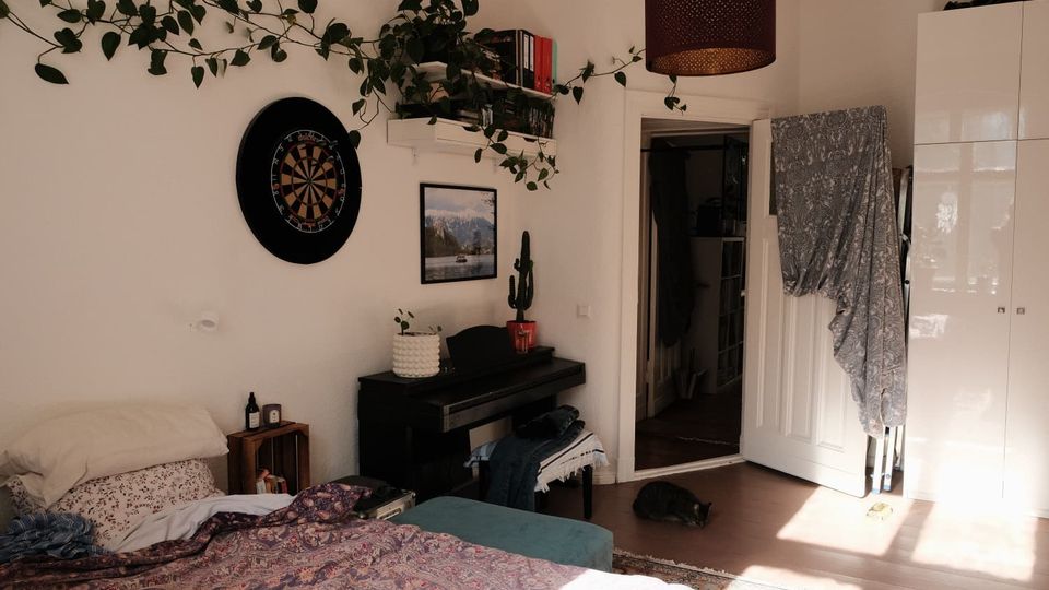 Helles Schlafzimmer mit Bett im Vordergrund, einem schwarzen Klavier an der Wand sowie Wandregalen mit Büchern und rankenden Zimmerpflanzen. Neben einer offenen Tür stehen ein großer weißer Kleiderschrank und ein Deckenlampenschirm; an der Wand hängen Dartscheibe und Bild, Sonnenlicht fällt auf den Holzboden.