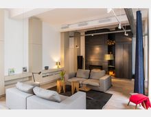 Modernes Wohnzimmer mit hellgrauen Sofas, einem schwarzen Teppich und kleinen Holztischen. Im Hintergrund sind dunkle Einbauschränke mit einem Kamin und Holzlager, sowie helle Wände mit Regalen und minimaler Dekoration sichtbar.