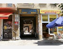 Eine schmale Gasse zwischen zwei Gebäuden, flankiert von einem Bäckereigeschäft mit Sitzmöglichkeiten im Freien auf der rechten Seite und einem Eingang mit Graffiti auf der linken Seite. Im Hintergrund ist ein Innenhof mit Personen und geparkten Autos sichtbar.