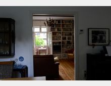 Wohnzimmer mit Holzboden, einer Bücherwand und einem Kronleuchter. Im Hintergrund Blick auf eine Veranda mit einem Holztisch und viel Tageslicht.