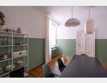 Esszimmer mit schlichtem Design, ausgestattet mit einem schwarzen Tisch und unterschiedlich farbigen Stühlen. Die Wände sind teils weiß, teils in Grün gestrichen, mit einem weißen Regal an der linken Wand, Holzboden und einer weißen Tür rechts im Raum.