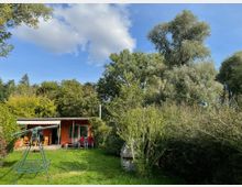 Ein kleiner Garten mit Rasen und Bäumen, in dessen Hintergrund eine orangefarbene Gartenhütte mit überdachter Terrasse steht. Im Vordergrund ist eine freistehende, grüne Schaukel sichtbar, umgeben von dichter Vegetation.