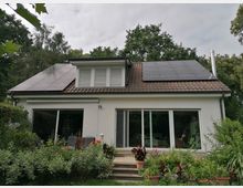 Einfamilienhaus mit weißer Fassade und einem Satteldach, auf dem Solarpaneele installiert sind. Im Vordergrund befindet sich ein Garten mit zahlreichen Pflanzen und Blumen, vor einer Terrasse mit Fenstertüren.