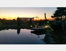 Ein Garten mit einem kleinen Teich im Vordergrund, einer Holzplattform am Ufer, und vereinzelten Lampen, die warmes Licht abgeben. Im Hintergrund ist eine flache Gebäude-Silhouette sichtbar, während die untergehende Sonne einen orangefarbenen Himmel erzeugt.