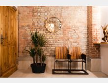 Raum mit einer Ziegelsteinwand und hellen Bodenfliesen, ausgestattet mit zwei hölzernen Stühlen, einer Topfpflanze, einer Holztür auf der linken Seite und einer dekorativen Lampe mit geometrischem Muster an der Wand.