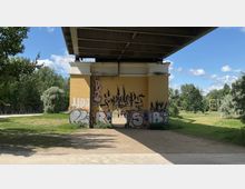 Unterseite einer Brücke mit einer gelben gemauerten Säule, die mit Graffiti bedeckt ist. Im Hintergrund befindet sich ein Park mit Bäumen und Wiesen unter einem blauen Himmel mit einigen Wolken.