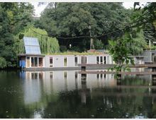 Ein modernes Hausboot mit großen Fenstern und einer Solaranlage auf dem Dach liegt auf einem ruhigen Gewässer, umgeben von dichter Vegetation und Bäumen. Im Vordergrund spiegelt sich das Hausboot im Wasser, während Zweige und Blätter einen Teil des Bildes einrahmen.