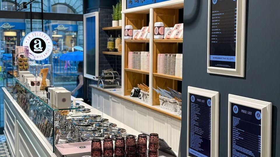 Ein moderner Eissalon mit einer langen Theke aus weißem Holz, darauf Metallbehälter für Eis, gestapelte Gläser und kleine Flaschen. Regale im Hintergrund enthalten Eisbecher und Zubehör, während digitale Preistafeln an der dunkelblauen Wand hängen. Durch ein großes Fenster ist eine Straße mit Fußgängern sichtbar.