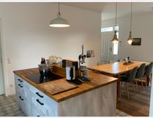 Moderne Wohnküche mit einer Kücheninsel aus Holz und weißen Schubladen, ausgestattet mit einer Herdplatte und Küchengeräten. Im Hintergrund befindet sich ein großer Esstisch aus Holz mit grauen Stühlen und hängenden Lampen an der Decke.