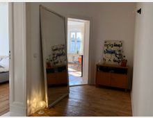 Ein Flur mit Holzboden, einer großen, angelehnten Spiegelplatte und einem kleinen Holzschrank mit Dekorationsgegenständen darauf. Im Hintergrund ist ein heller Raum mit einem Fenster und einem Tisch mit Stühlen erkennbar.