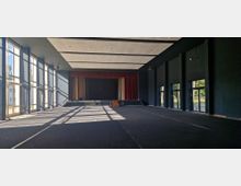 Großer, dunkler Veranstaltungssaal mit einer Bühne im Hintergrund, umrahmt von roten Vorhängen. Links befinden sich hohe Fenster, die Sonnenlicht in den Raum lassen, und rechts sind mehrere verglaste Türen sichtbar.