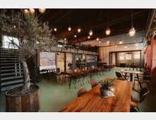 Kaffeebar oder Bistro in industriellem Stil mit Holztischen, modernen Stühlen und einem großen Olivenbaum in einem runden Holztopf. Die Einrichtung umfasst hängende Metalllampen, eine Treppe mit schwarzem Geländer sowie eine Galerieebene, und die Wände sind dekoriert mit Bildern und Glastüren.