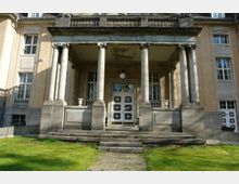 Neoklassizistischer Eingangsbereich eines historischen Gebäudes mit symmetrischer Anordnung von Säulen, einer verzierten weißen Tür und umliegender Grünfläche. Die Fassade ist aus beigefarbenem Stein mit großen Fenstern.