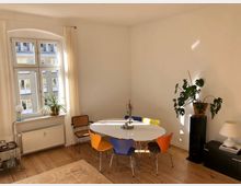 Helles Wohnzimmer mit Holzdielenboden, einem großen Fenster mit Vorhängen und einer weißen Heizung darunter. Ein runder, weißer Esstisch mit bunten Stühlen steht in der Ecke, daneben Pflanzen auf einem Regal und auf der Fensterbank.