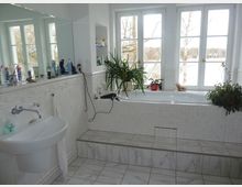 Helles Badezimmer mit weißen Fliesen, einem eingebauten Waschbecken links und einer erhöhten Badewanne rechts, die von mehreren Zimmerpflanzen dekoriert ist. Große Fenster im Hintergrund lassen Tageslicht in den Raum und geben den Blick auf eine baumreiche Außenlandschaft frei.