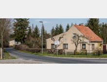 Straßenszene mit einem beige gestrichenen Einfamilienhaus mit rotem Ziegeldach und umzäuntem Garten, umgeben von kahlen Bäumen und einer niedrigen Hecke. Im Hintergrund stehen Nadelbäume, und ein schmaler asphaltierter Weg führt vorbei.