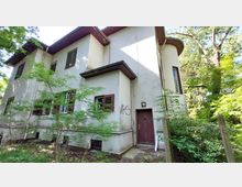 Zweistöckiges Wohnhaus mit hellgrauer Putzfassade, brauen Fensterläden und einer braunen Eingangstür, umgeben von Bäumen und wildem Grün. An der Wand ist Graffiti erkennbar, und das Gebäude wirkt von Vegetation teils verdeckt.