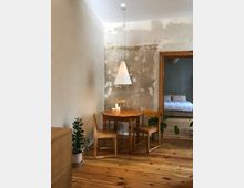 Ein schlichter, rustikaler Innenraum mit einem runden Holztisch, zwei Holzstühlen und einer hängenden weißen Pendelleuchte. Die Wände haben eine teilweise unverputzte Optik, der Boden besteht aus hellen Holzdielen, und im Hintergrund ist ein Schlafzimmer mit einem weißen Bett sichtbar.