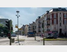 Straßenecke in einer Stadt mit mehrgeschossigen Wohnhäusern im Altbaustil, Verkehrsampeln und Straßenschildern. Im Hintergrund ist eine U-Bahn-Station mit dem Schild „Friedrich-Wilhelm-Platz“ zu sehen, daneben Geschäfte im Erdgeschoss der Gebäude.