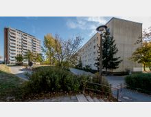 Mehrstöckige Wohngebäude in einer urbanen Umgebung, umgeben von Gehwegen, Bäumen und Büschen. Im Vordergrund führen Treppenstufen zu einem Weg hinunter, der zwischen den Gebäuden verläuft.