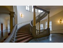 Großes, elegantes Treppenhaus in einem historischen Gebäude mit geschwungener Treppe, verzierten Geländern und Säulen. Die Wände sind beige, mit goldenen Wandlampen und hohen Fenstern, die natürliches Licht einfallen lassen.