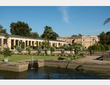 Ein historisches Gebäude mit Arkaden und großen Fenstern, umgeben von einem gepflegten Garten mit Palmen, Blumenbeeten und einer Balustrade. Im Vordergrund befindet sich ein steinerner Brunnen mit einem kleinen Wasserstrahl.