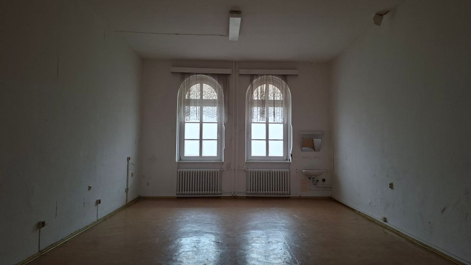 Leerer, schlicht gehaltener Innenraum mit weißen Wänden und braunem Boden, beleuchtet durch zwei große Rundbogenfenster an der Stirnwand. Unter den Fenstern stehen Heizkörper; rechts ist ein kleines Wandwaschbecken mit Ablage und sichtbaren Anschlüssen montiert.