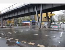 Straßenszene mit einer nassen Fahrbahn unter einer erhöhten Eisenbahnbrücke aus Stahl, daneben ein Gebäude mit der Aufschrift "Hähnchenhaus". Im Hintergrund stehen weitere Gebäude und einige Bäume, Verkehrszeichen zeigen die Richtung zum Tiergarten.