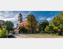 Historische Kirche mit einem hohen Glockenturm und einem roten Ziegeldach, umgeben von Bäumen und einer Kopfsteinpflasterfläche im Vordergrund. Der Himmel ist klar mit einigen Wolken, und die Umgebung zeigt eine ruhige, parkähnliche Landschaft.