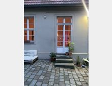 Hinterhofansicht mit gepflastertem Boden, einer cremefarbenen Fassade und einer Glastür, die von zwei Fenstern flankiert wird. Pflanzen in Töpfen stehen neben einer kurzen Treppe, und links ist eine Sitzbank aus Holzpaletten mit Kissen zu sehen.