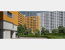 Mehrstöckige Wohngebäude, darunter ein gelbes Gebäude mit Balkonen und ein weißes Hochhaus im Hintergrund. Vor den Gebäuden befinden sich grüne Bäume und Sträucher sowie eine gepflasterte Fläche, auf der eine Person mit einem Rollkoffer zu sehen ist.