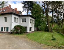 Zweistöckiges Wohnhaus mit weißer Fassade, rotem Satteldach und grünen Fensterläden, umgeben von einer Grünfläche mit Bäumen und einem gepflasterten Weg im Vordergrund. Im Hintergrund stehen hohe Bäume, die den Garten einrahmen.