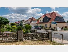 Wohnsiedlung mit Einfamilienhäusern in Reihenbauweise, gekennzeichnet durch rote Ziegeldächer und helle Fassaden. Im Vordergrund ein gepflegter Garten mit Ziersträuchern und einer niedrigen Betonmauer; daneben eine Straßenecke mit Straßenschild.