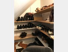 Schuhregal in einem kleinen Raum mit schräger Decke. Mehrere Holzregale sind mit verschiedenen Schuhen, darunter Stiefel, High Heels und Sportschuhe, sowie einer Kartonschachtel gefüllt.