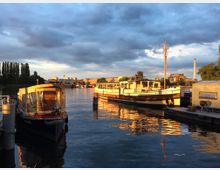 Hafenansicht bei Sonnenuntergang mit zwei Booten am Wasser, darunter ein größeres weißes Boot mit dem Namen "ARCONA". Im Hintergrund sind moderne Gebäude, Bäume und ein Schornstein zu sehen, während das Wasser die Farben des Himmels reflektiert.